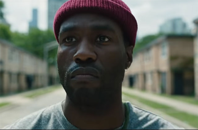 Yahya Abdul-Mateen II. Black man wearing dark red wool skull cap and green t-shirt standing in front of a row of buildings.