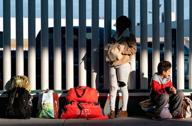 A parent and child hold all their belongings and lean against a large, white fence.
