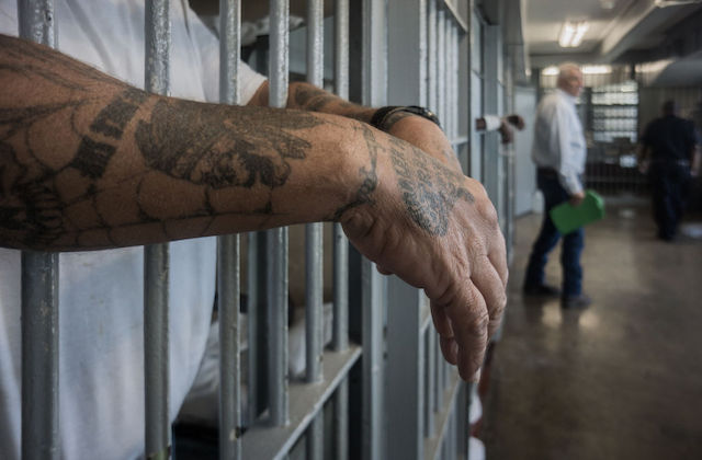 a man with tattoos hangs his arms through the bars of a prison cell.