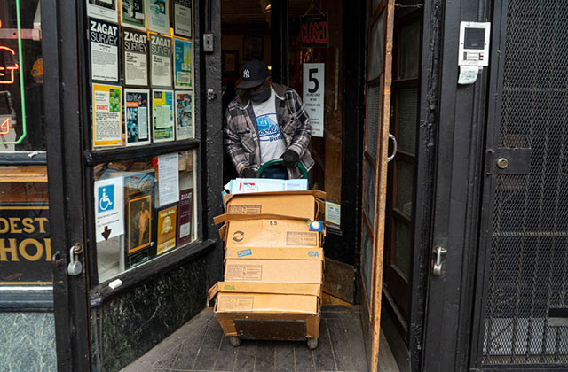 COVID-19 NYC 2020. Black man wearing dark Yankees cap , T-shirt and plaid jacket pulling boxes into the doorway of a shop.