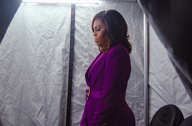 Michelle Obama. Black woman with dark shoulder length hair standing in a concentrated lit space wearing a purple suit.