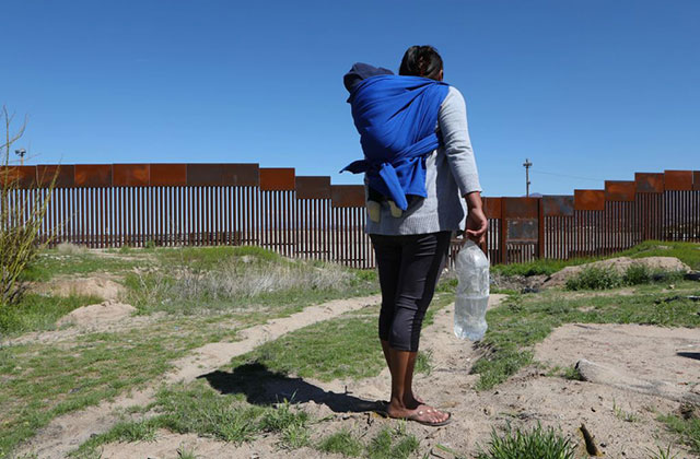 Migrant. Rear view of woman with dark hair wearing gray top, dark leggings and carrying a child in a blue blanket and a large bottle of water, facing the U.S./Mexico border wall.