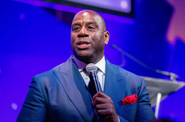 Magic Johnson. Black man with bald head wearing blue suit jacket, white shirt with red pocket square, holding a mic.