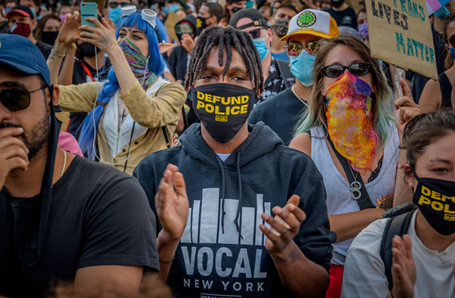 Defund Police. Black male protestor in a crowd wearing a black face mask that reads "defund police" in yellow letters, wearing a black hoodie.