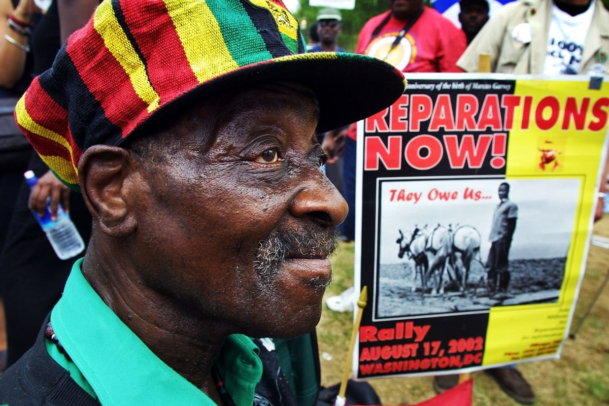 Older Black man with a red, black, green, and gold "rasta cap" with brim, holding a "Repaations Now!" sign
