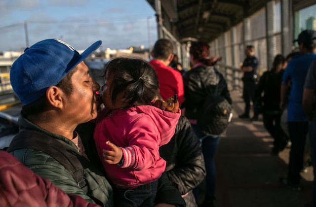 A man wearing a blue baseball cap kisses his small daughter as he holds her in his arms.