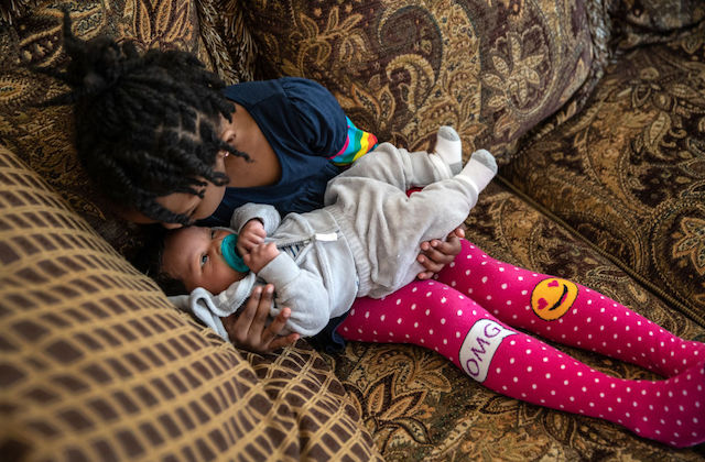 A little Black girl wearing pink stockings sits on a couch and kisses her newborn sibling on the head