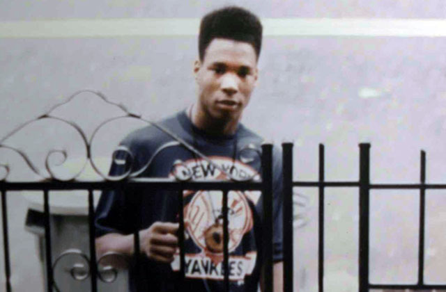 Yusuf Hawkins. Black teenage boy with high top wearing dark tee standing in front of a gate.