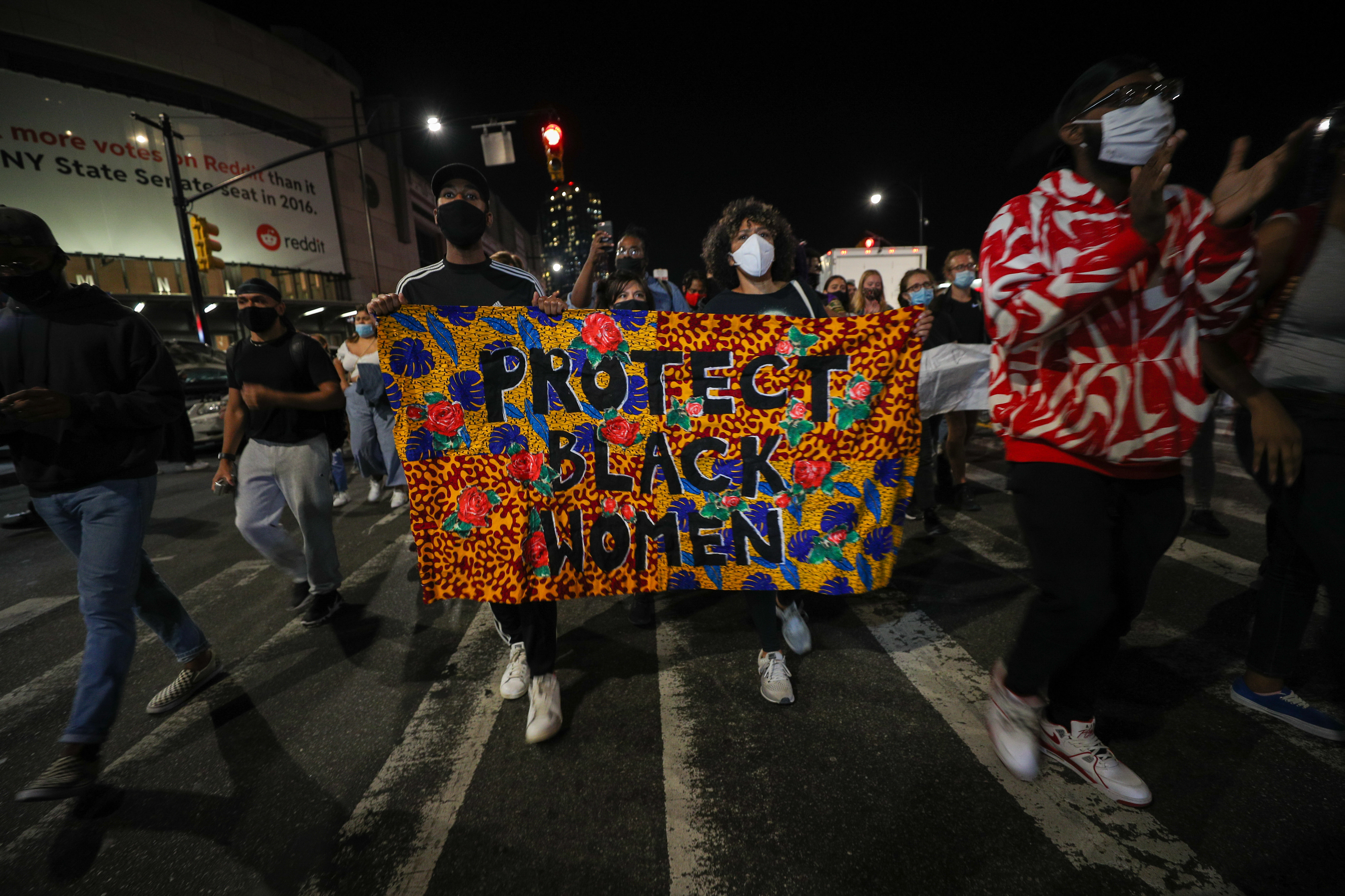 People march at night, carrying a colorful banner saying "Protect Black Women."