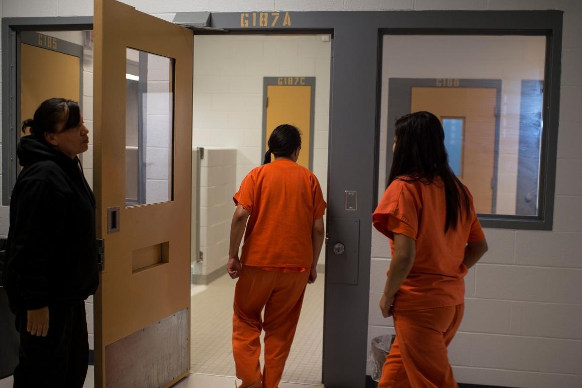 Two young adults dressed in orange jail suits go through a door as a woman guard holds it open for them
