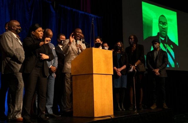About 20 people onstage with a image of George Floyd on a screen behind them. There is a podium and microphone. Press Conference