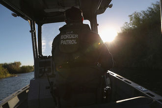 Border Patrol stands and rides small boat near U.S. Mexico border> His back is to the camera and the back of his jacket reads, Border Patrol. 