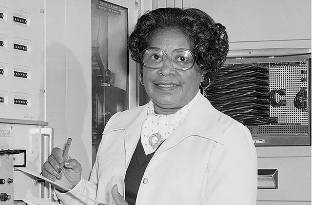 Mary Jackson. Black and white photo of Black woman with dark hair wearing white lab coat and glasses, holding a notepad and pen.
