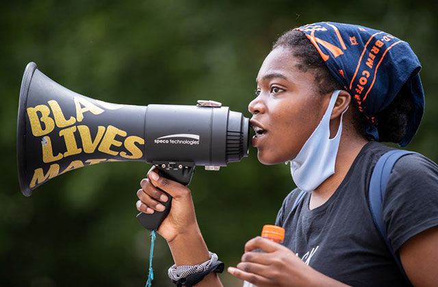 BLM-protest. Young Black woman wearing blue and orange headkerchief, white face mask around her chin and black Tee holding black bullhorn reading Black Lives Matter in yellow words.