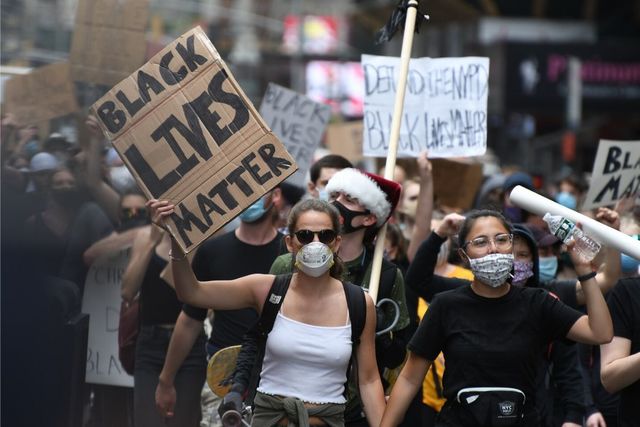 Protest photo with two white women at the front both with masks on, one holding a sign saying, "Black Lives Matter" 