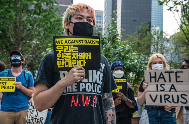 Stop AAPI Hate. Asian man with colorful hair wearing black mask and NWA Tee holding a sign reading "We against racism."