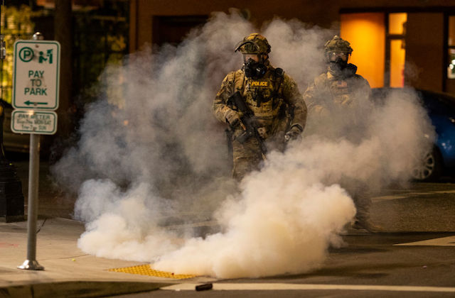 A federal officer wearing military gear walks in a city street surrounded by tear gas.