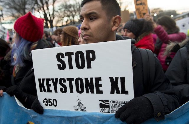 A shot of a young brown man standing outside at a protest and holding a sign that reads "Stop Keystone XL."