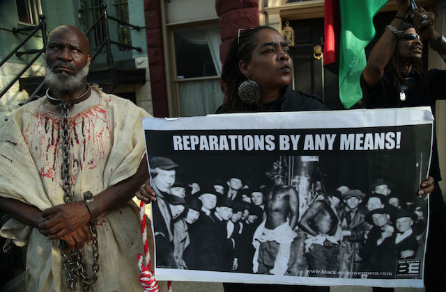 Three Black people hold a large protest sign that reads "Reparations By Any Means!"
