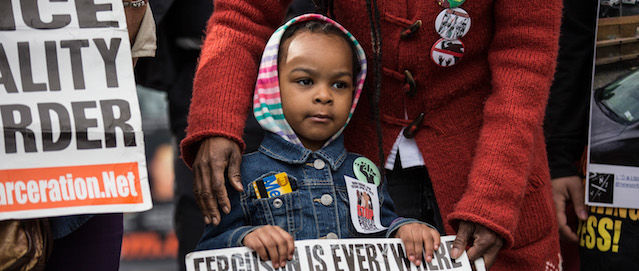 A little Black girl in a striped hoodie holds up a sign during a protest