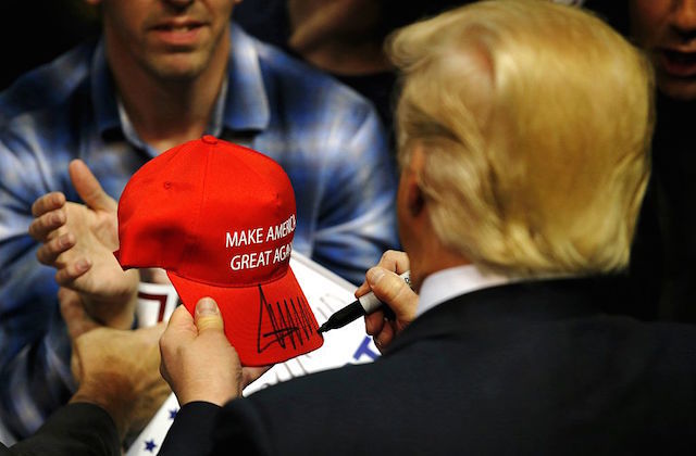 Man stands with back to camera holding a red hat that says "Make American Great Again"; man in blue plaid shirt holds out hands to receive the hat