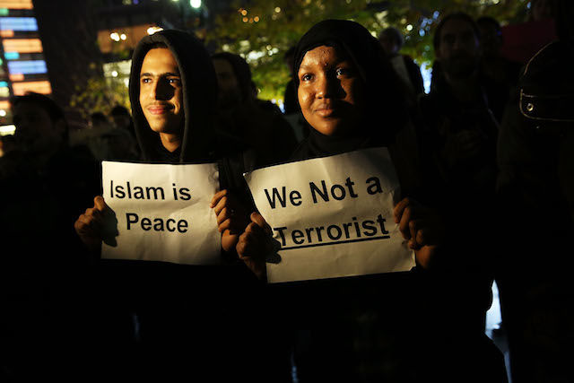 A young Muslim man wearing a black hoodie and a young woman wearing a hijab hold up signs that say "Islam is peace" and "We not a terrorist"