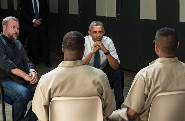 White man with navy shirt and blue jeans next to Black man in white shirt and navy tie and navy pants in front of men in brown prison uniforms, all against grey floor and white wall with blue doors