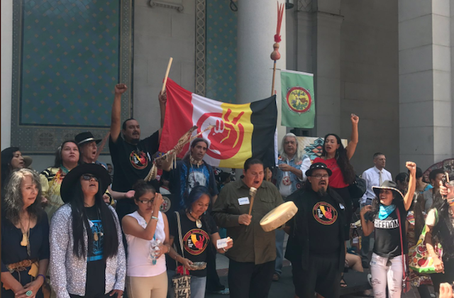 Brown people in multicolored clothing stand waving green and red-white-yellow-and-black flags on steps of building with green and white stone