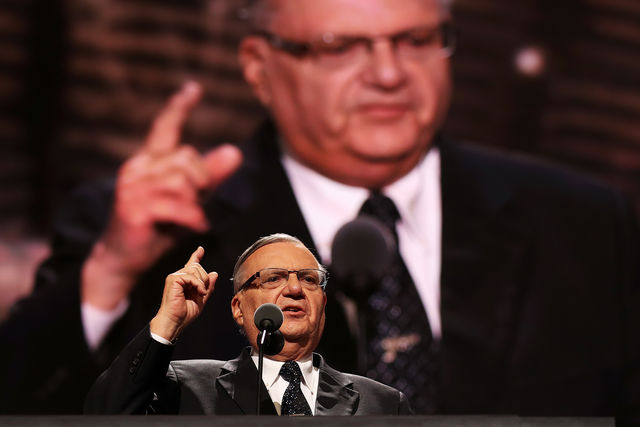 Joe Arpaio stands at a podium and gestures with his index finger. An image of himself is blown up on a screen behind him. 