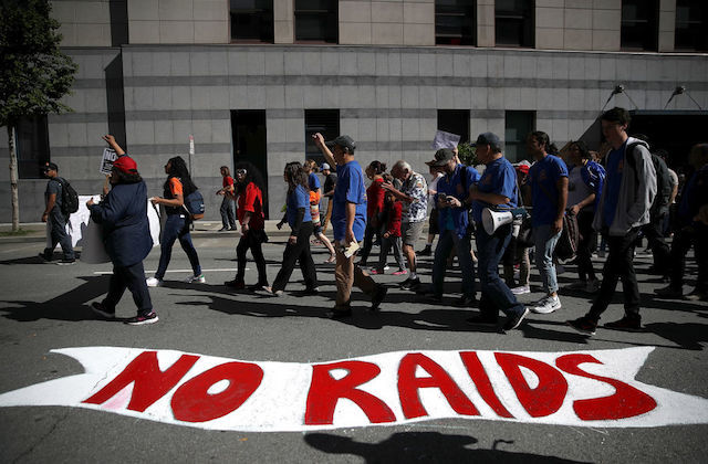People in blue t-shirts and multicolored clothing walk on grey street with red and white spraypaint image in front of grey buildings