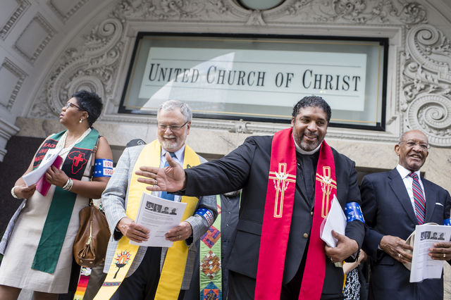 Four faith leaders holding papers standing in front of United Church of Christ.