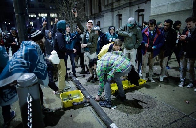 White men and women in green and and black and white and grey clothing stand over grey and yellow traffic pole on grey sidewalk in front of grey buildings and black night sky