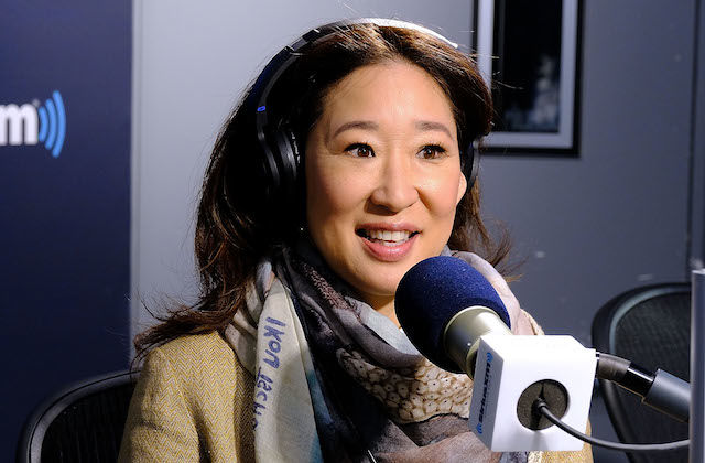 Asian woman with in brown blazer and scarf with blue text and black headphones behind blue and white microphone and black stand in front of navy and grey walls