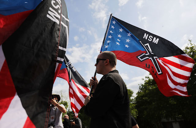 A white man holds a red white blue and black flag with white supremacist symbols and the acronym "USA"
