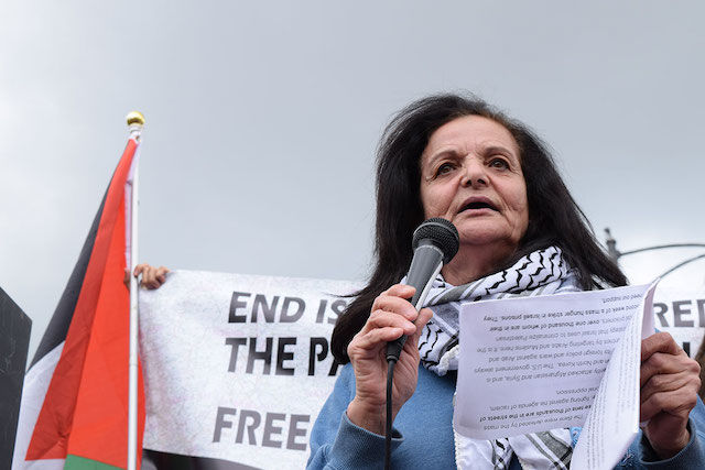 A Palestinian woman wearing a keffiyeh around her neck speaks to a crowd
