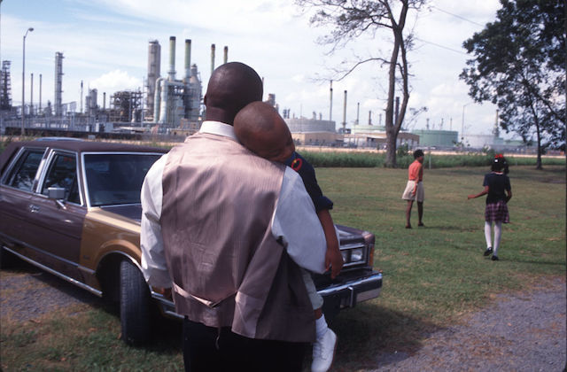 A Black man holds a Black boy standing in front of chemical plants