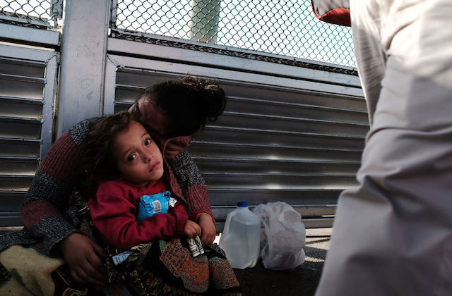 Small girl with brown hair and wearing a red t-shirt sits in her mother's lap in front of a grated fence. 