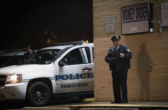 White police officer stands in front of a store at night with police car visible in the background.