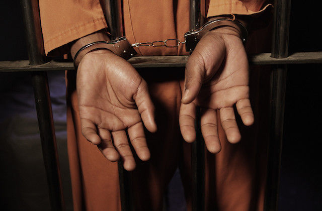 Black hands poke through orange sleeves, handcuffed around metal bars
