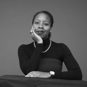 Toccara Castleman in a black and blue, sleeved dress with head resting in one hand against a red background.