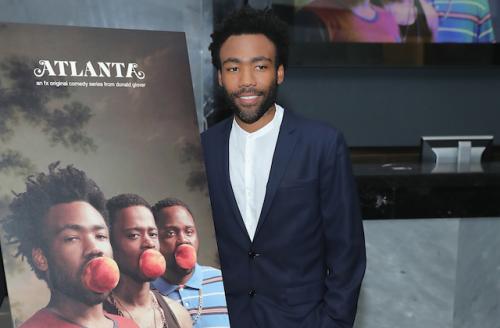 Donald Glover in navy suit with white shirt next to sign on black easel