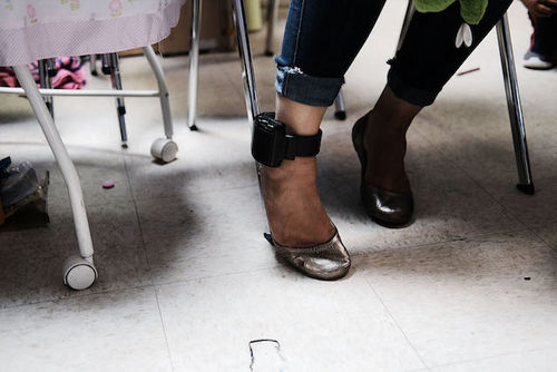 A brown woman wearing metallic gold heels shows her electronic monitoring bracelet.