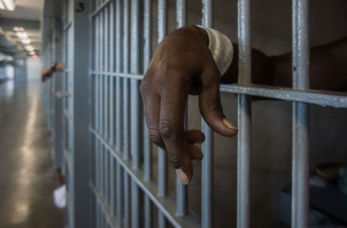 Black person's hand shown through prison cell bars.