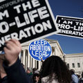 Two protestors holding black signs with white font that say "i am the pro-life generation" attempt to block a blue sign with white font that says "keep abortion legal" at a rally outside the supreme court