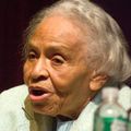 Black woman with grey hair in green shirt speaks into black microphone in front of brown background