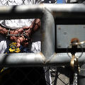 A person with their hands shackled behind their back is seen through an opening in a chain-link fence