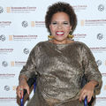Ntozake Shange. Black woman with short, curly brown hair smiles in gold dress and blue wheelchair in front of white screen with brown and yellow logos and text