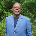 Henry Louis Gates Jr. A Black man with a blue jacket and gray beard stands outside with green lawn behind him.