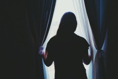 A dark-lit image of a woman with her back to the camera as she looks out the window