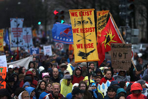 Brown woman in black jacket holds yellow sign with red text and black images in crowd of Brown and Black and White people in multicolored clothing with multicolored signs on grey pavement